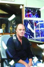 A man in a dark shirt sits in a chair in front of colorful abstract artwork.