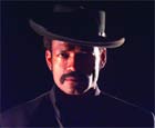 A man with a mustache wearing a black wide-brimmed hat and turtleneck against a dark background.