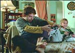 A man and a young child sit on a couch playing video games in a living room.