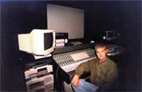 A man sits in a dark studio surrounded by monitors, a mixing console, and audio equipment.