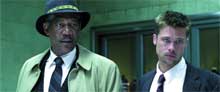 A man in a fedora and tan coat stands next to a man in a dark suit.