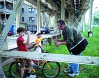 A man leans toward a young boy sitting on a bicycle under a metal bridge structure.