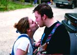Two people leaning in to kiss while holding musical instruments outdoors.