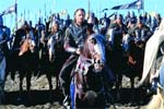 A man in dark clothing rides a horse at the front of a large group of mounted riders under a blue sky.