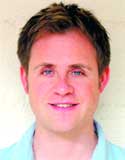 A smiling man with short brown hair and blue eyes wearing a light blue collared shirt.