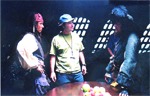 Three men stand in a dark room near a table, two wearing costumes and one in casual clothing.