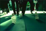 Small clear glass bottles sit on a teal floor as people walk past in a blurred background.