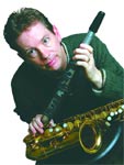A man looking at the camera while holding a flute vertically and a saxophone horizontally.
