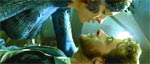 A woman in a blue dress leans over a man with a beard who is lying on his back.