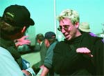 A blonde man in a black shirt talks to a person wearing a black baseball cap.