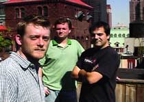 Three men posing together on a rooftop with a brick building in the background.