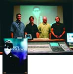 Four men stand behind a large audio mixing console in a dark studio with a projection screen behind them.
