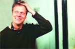 A smiling man in a dark long-sleeved shirt rests his hand on his head against a light background.
