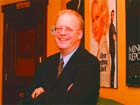A smiling man with glasses wearing a dark suit and tie, standing in a room with posters on the wall.