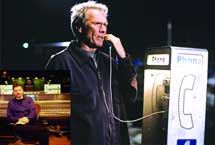 A man stands next to a white public telephone booth, holding the receiver to his ear.