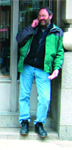 A man with a beard wearing a green jacket, black shirt, and blue jeans stands leaning against a building.