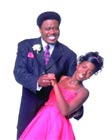 A man in a black suit and a woman in a pink dress dancing and smiling against a white background.
