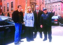 Four people standing on a city sidewalk in front of brick buildings and a black car.