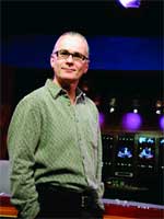 A man wearing glasses and a green button-down shirt stands in a dark studio with monitors in the background.