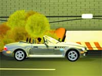 A silver convertible car driving along a road past yellow trees and a brick wall.