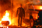 A man in a dark jacket and sunglasses stands amidst fire and a burning pickup truck.
