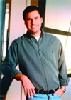A smiling man wearing a dark green button-down shirt and blue jeans, standing with one hand on his hip.