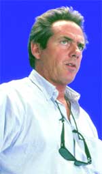 A man with short brown hair wearing a light blue button-down shirt against a solid blue background.