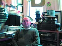 A man in a dark sweater sits in a room filled with electronics, books, and various objects.