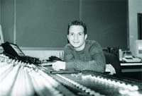 A man smiling while sitting behind a large audio mixing console in a recording studio.