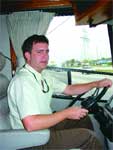 A man in a light-colored shirt sits in a vehicle's driver seat with his hands on the steering wheel.