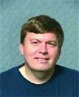 A man with short brown hair smiling, wearing a dark blue shirt against a textured blue background.