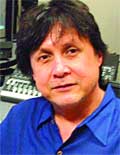 A man with dark hair wearing a blue collared shirt looking forward.