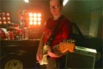 A person in a red shirt plays an electric guitar in a dark room with bright stage lights in the background.