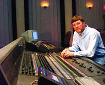 A man in a light blue shirt sits behind a large professional audio mixing console in a dimly lit studio.