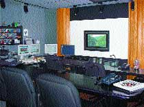 A dark control room with multiple computer monitors, a large screen on a white wall, and black office chairs.