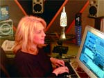 A blonde woman sits at a desk using a computer in a room with studio speakers and equipment.