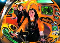 Two people wearing harnesses sit inside a brightly lit, orange-colored amusement park ride with glowing green controls.