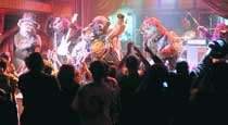 Costumed performers play instruments on a brightly lit stage for a crowd of people in silhouette.