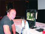 A smiling man in a black shirt sits at a table with two irons and a computer monitor.