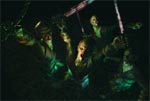 Performers in dark costumes with dramatic green and white lighting in a dark setting.