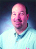 A smiling man with short dark hair and a goatee wearing a light blue button-down shirt.