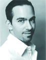 A headshot of a man with dark hair and a goatee wearing a white collared shirt.