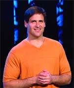 A smiling man with short dark hair wearing an orange v-neck t-shirt against a dark blue background.