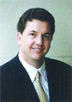 A man with dark hair smiling, wearing a dark suit, white shirt, and patterned gold tie.