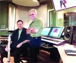 Two men standing in a music studio with computer monitors, speakers, and a guitar.
