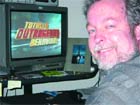 A smiling man sits in front of a television screen displaying colorful text.