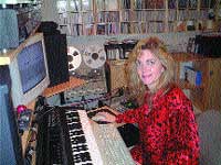 A woman in a red patterned shirt sits at a desk with a computer monitor, keyboard, and audio equipment.