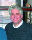 A smiling man with short gray hair wearing a dark gray sweater over a white collared shirt.