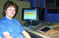 A man in a blue shirt sits in front of a computer monitor and a large audio mixing console.