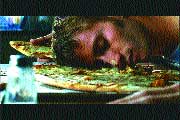 A person lies face down with their head resting on a large pizza on a table.